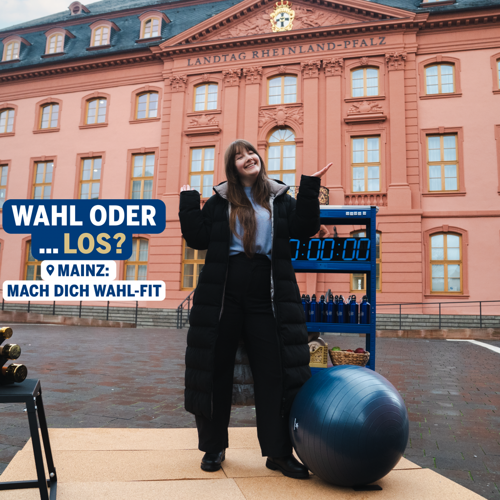 Eine junge Frau steht lächelnd auf einer kleinen Trainingsfläche vor dem Landtag von Rheinland-Pfalz in Mainz und hebt fragend die Hände. Neben ihr befinden sich Sportgeräte wie Hanteln, ein Gymnastikball und eine Trainingsuhr. Im Bild ist der Schriftzug zu sehen: „Wahl oder … los? Mainz: Mach dich wahl-fit“. Das Bild verbindet das Thema Wahl mit einem spielerischen Trainingsmotiv vor dem Parlamentsgebäude.