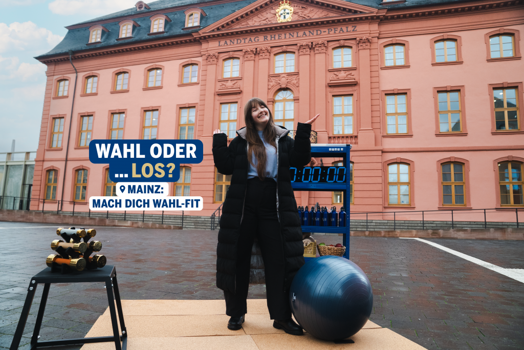 Eine junge Frau steht lächelnd auf einer kleinen Trainingsfläche vor dem Landtag von Rheinland-Pfalz in Mainz und hebt fragend die Hände. Neben ihr befinden sich Sportgeräte wie Hanteln, ein Gymnastikball und eine Trainingsuhr. Im Bild ist der Schriftzug zu sehen: „Wahl oder … los? Mainz: Mach dich wahl-fit“. Das Bild verbindet das Thema Wahl mit einem spielerischen Trainingsmotiv vor dem Parlamentsgebäude.