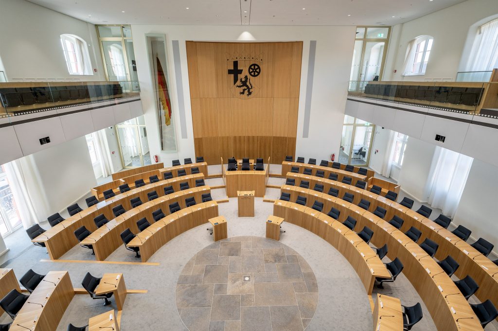 Blick in den Plenarsaal des Landtags Rheinland-Pfalz. Der Saal ist halbrund angelegt mit mehreren Reihen aus hellen Holztischen und schwarzen Sitzplätzen. In der Mitte befindet sich das Rednerpult, dahinter die Präsidiumsplätze. An der Stirnwand ist das Wappen von Rheinland-Pfalz angebracht. Der Raum ist hell, modern und symmetrisch gestaltet.