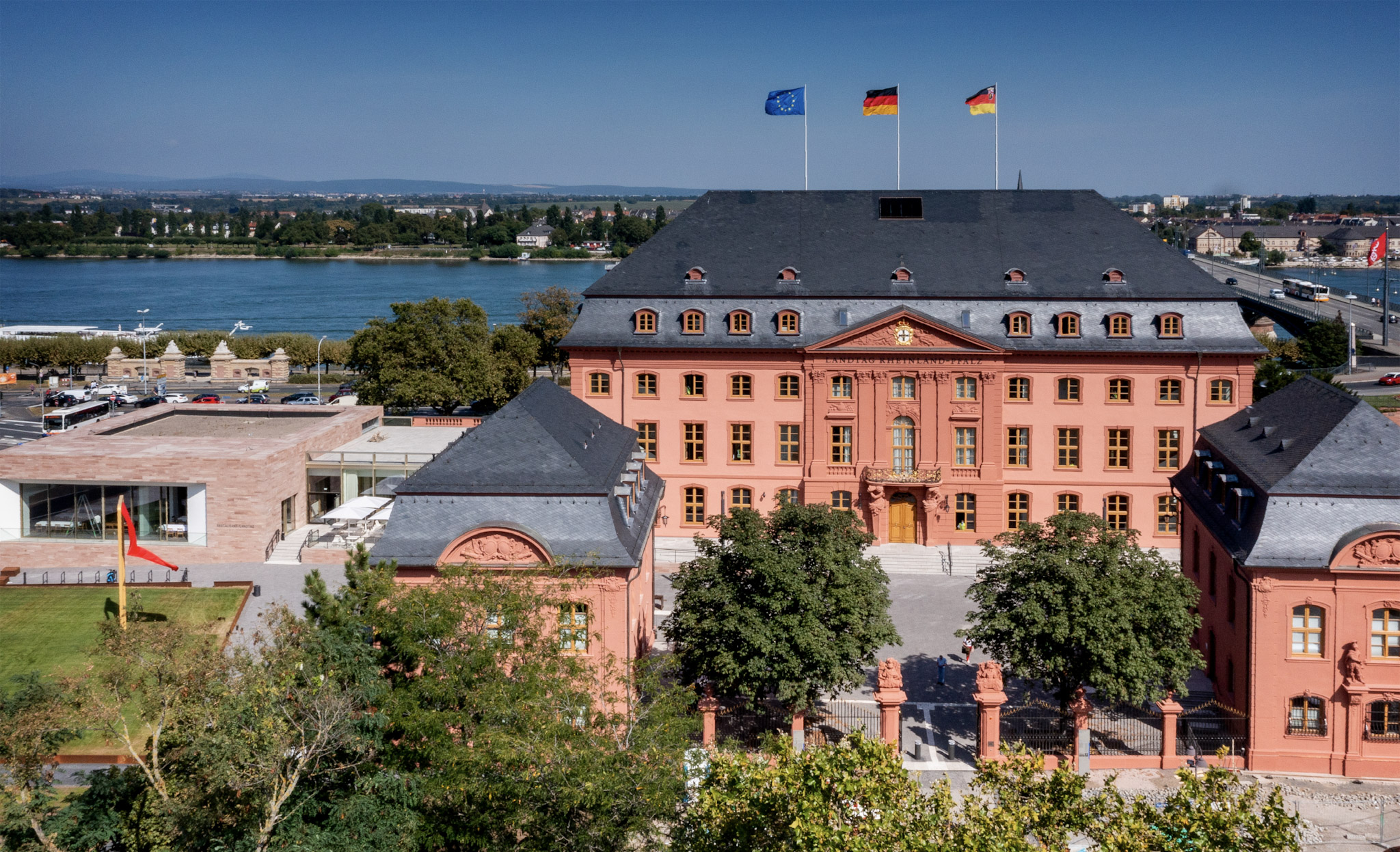 Luftaufnahme des Landtags von Rheinland-Pfalz in Mainz. Zu sehen ist das rosafarbene historische Parlamentsgebäude mit Innenhof und angrenzendem modernen Anbau. Auf dem Dach wehen die Flaggen der Europäischen Union, Deutschlands und von Rheinland-Pfalz. Im Hintergrund fließt der Rhein, daneben sind Straßen, Bäume und Teile der Stadt zu erkennen.
