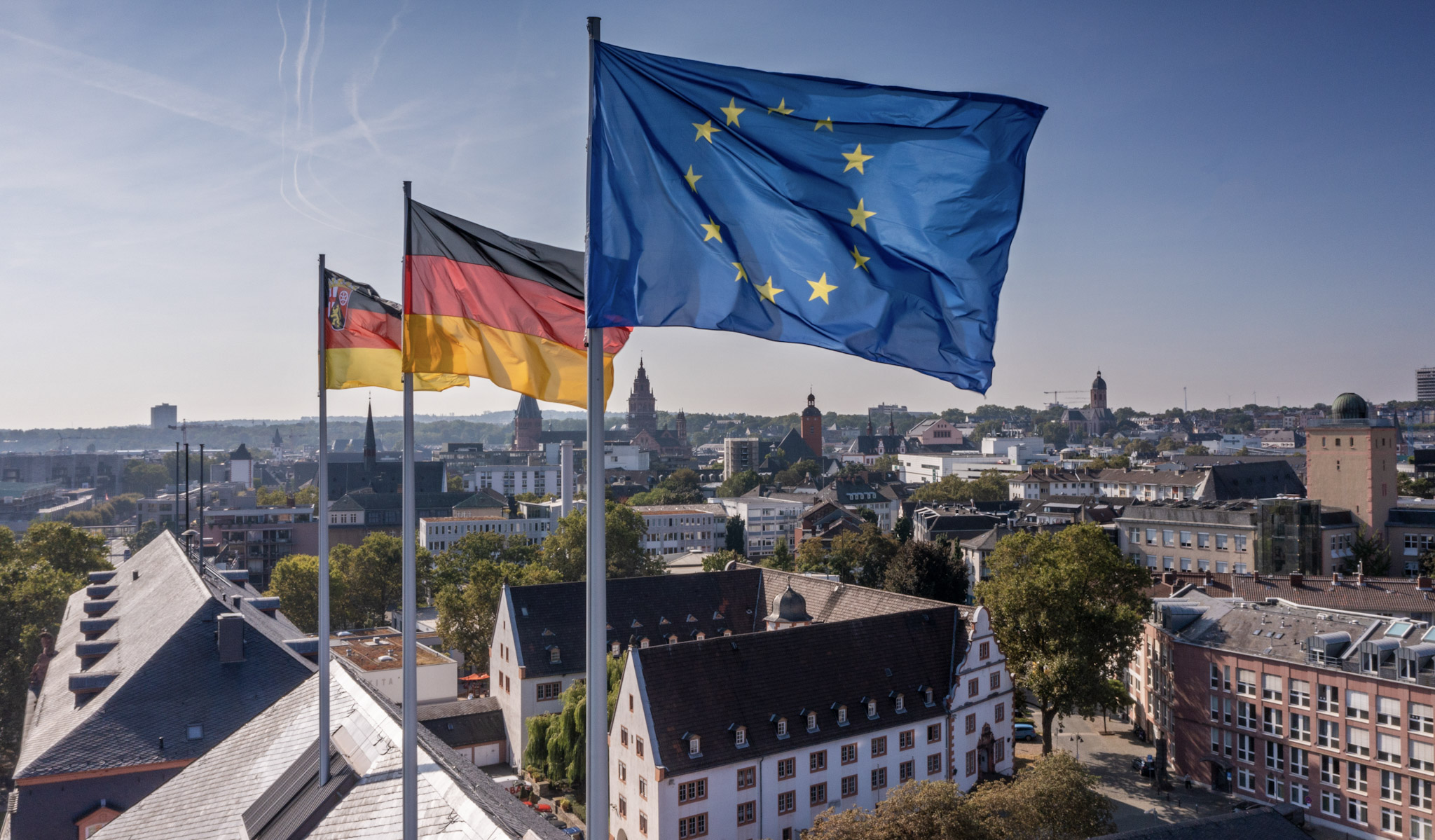 Blick über die Stadt Mainz mit historischen und modernen Gebäuden. Im Vordergrund wehen auf drei Fahnenmasten die Flaggen der Europäischen Union, Deutschlands und von Rheinland-Pfalz. Die Fahnen sind vor blauem Himmel zu sehen, im Hintergrund erstreckt sich die Stadtlandschaft mit Kirchtürmen und Wohnhäusern.