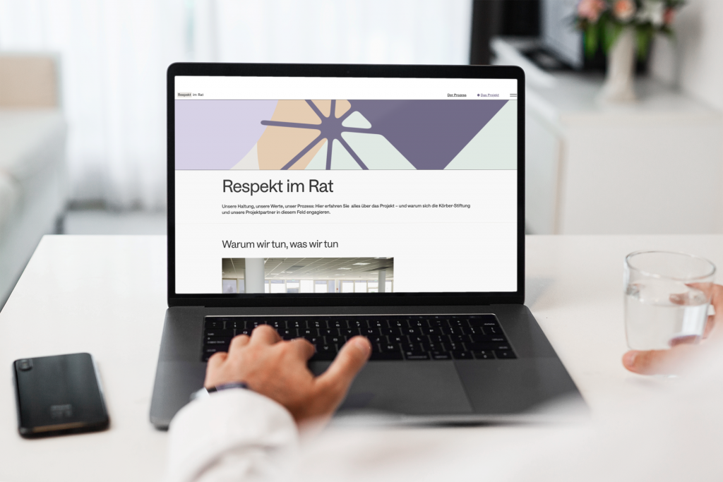 Ein Computer zeigt die Website von "Respekt im Rat" im Vordergrund sitz eine Person im weißen Hemd, wir schauen über die Schulter auf das Laptop auf welchem die Website "Respekt im Rat" aufgerufen ist. Das Bild ist hell und freundlich, der Raum um den Laptop zeigt viel Weiß und unterschiedliche Ecken in einem Raum.