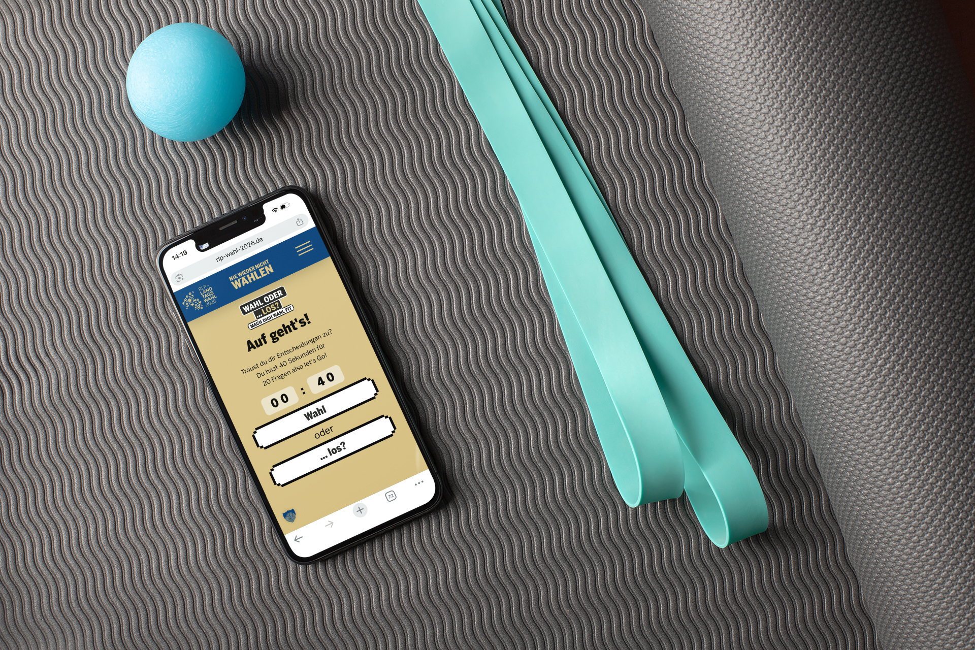 Smartphone liegt auf einer grauen Sport- oder Yogamatte. Auf dem Display ist die Website der Kampagne „Nie wieder nicht wählen“ mit dem digitalen Wahl-Training zu sehen. Daneben liegen ein türkisfarbenes Fitnessband und ein kleiner blauer Ball. Das Bild verbindet das Thema Wahl mit Bewegung und Training.